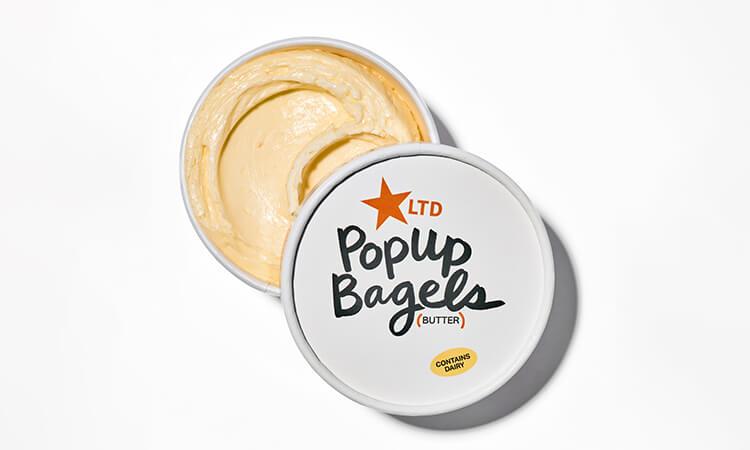 Open container of butter with "Popup Bagels" logo, revealing smooth, creamy texture. Lid marked with a red star and “Contains Dairy” label.